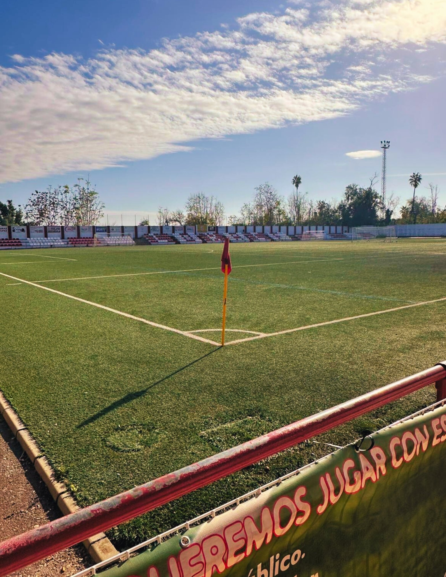 A scenic view of a pristine soccer field at Lora del Rio in Spain, featuring a red corner flag under a bright blue sky with scattered clouds.